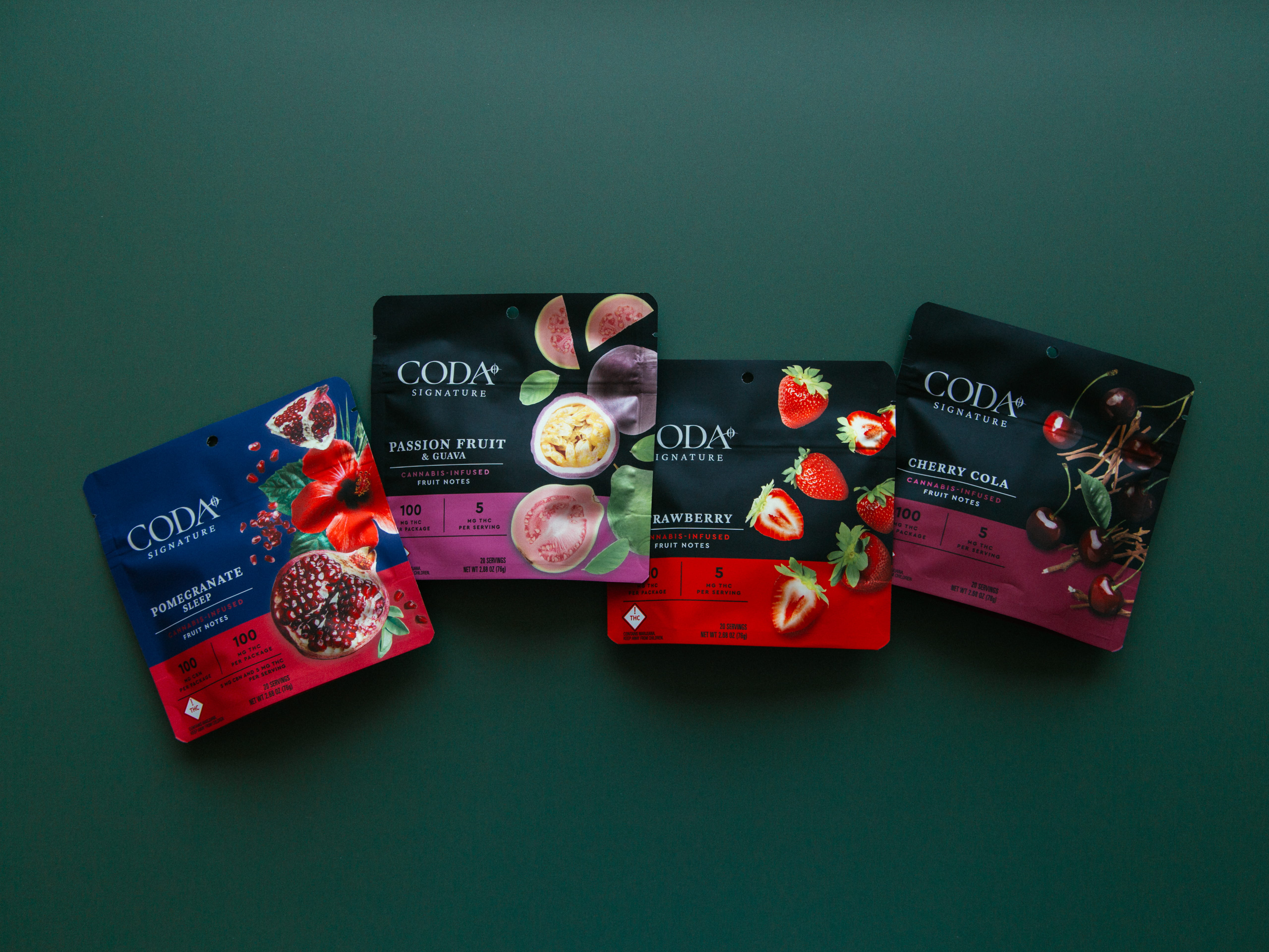 Four colorful CODA Signature cannabis-infused fruit notes packages with flavors Pomegranate, Passion Fruit & Guava, Strawberry, and Cherry Cola on a dark green background.