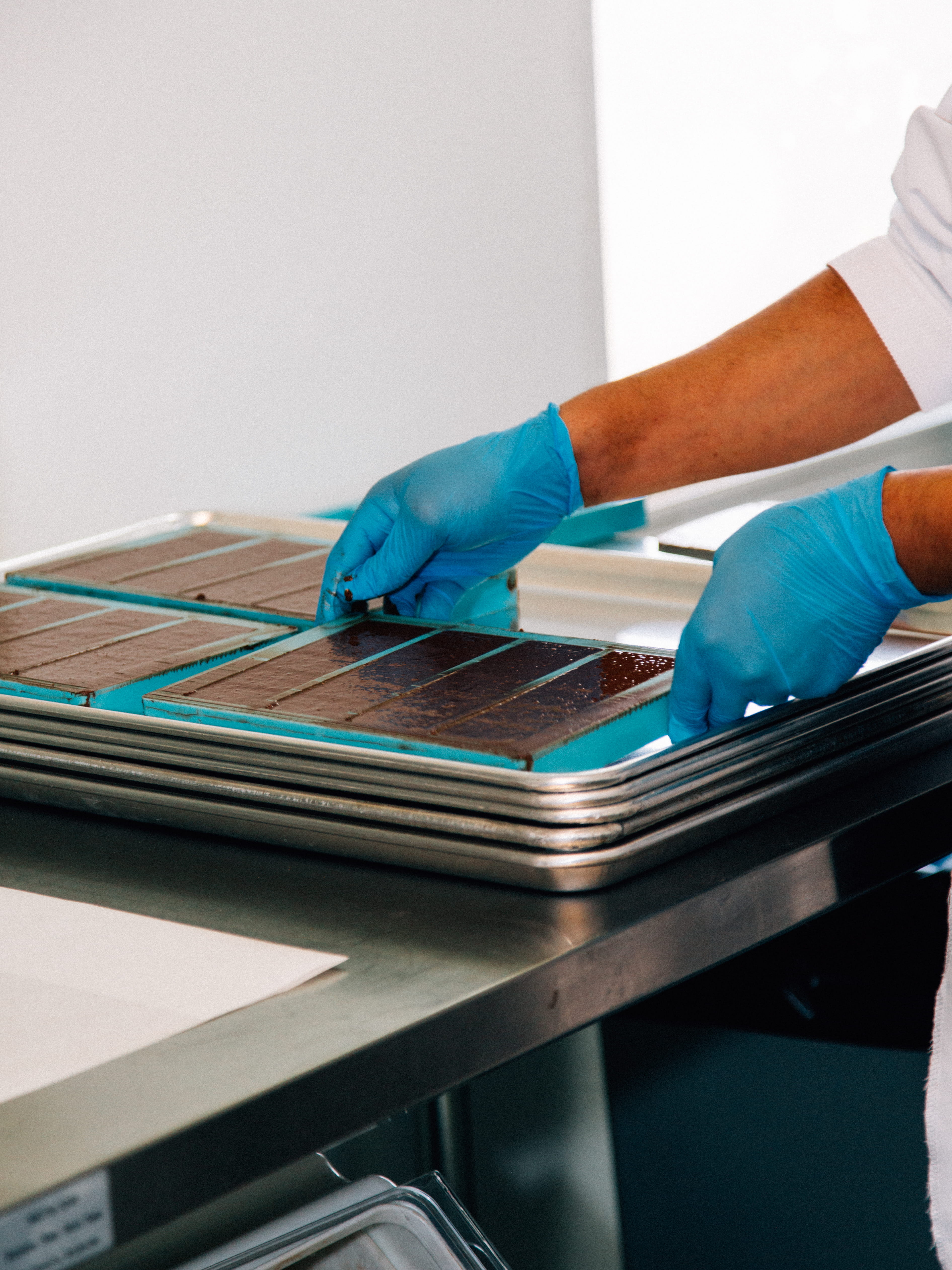 Person wearing blue gloves handling trays of chocolate bars on a metal table.