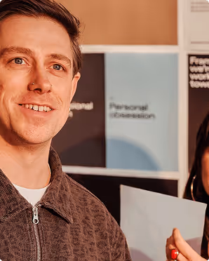Smiling man with short hair wearing a brown jacket and a nose ring, standing indoors near a bulletin board with blurred posters.