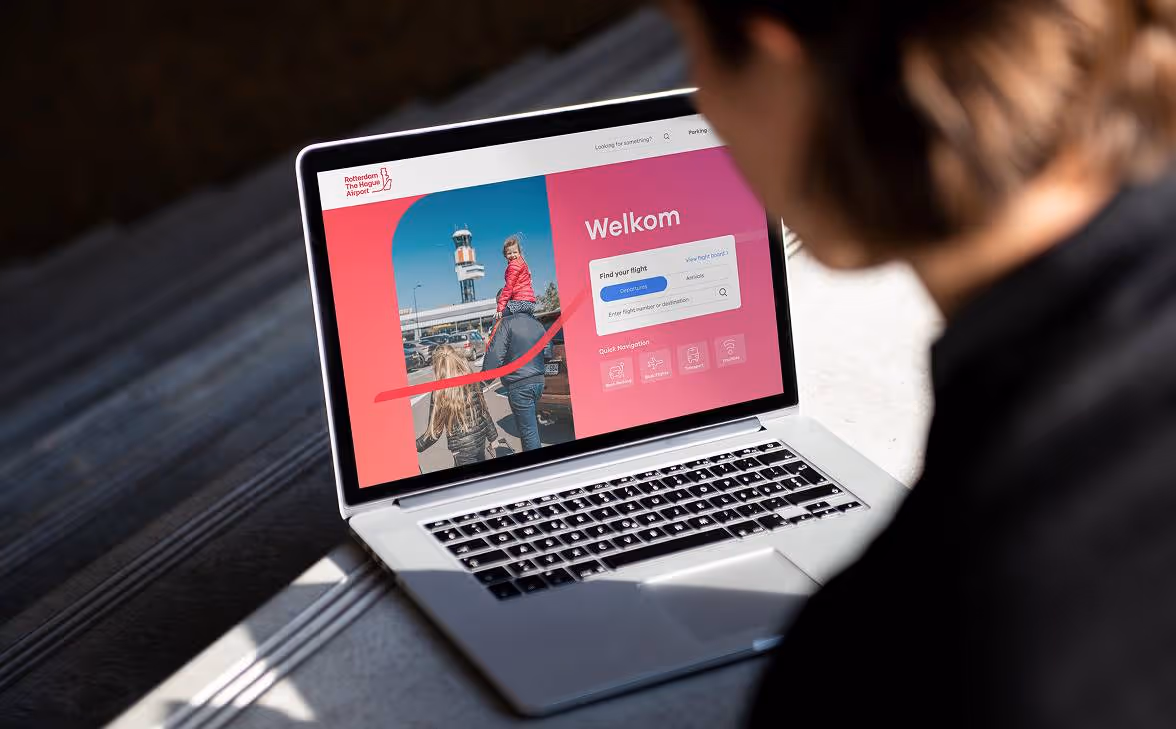 Person looking at a laptop screen showing the Rotterdam The Hague Airport welcome page with flight search options and a family photo.