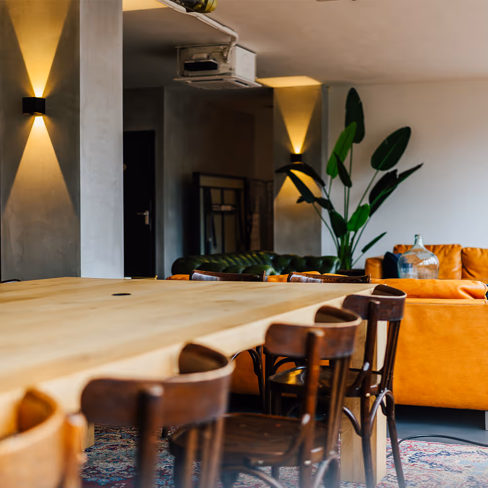 Modern office lounge area with a light wooden table, dark wooden chairs, leather couches, a large indoor plant, and warm wall lighting.
