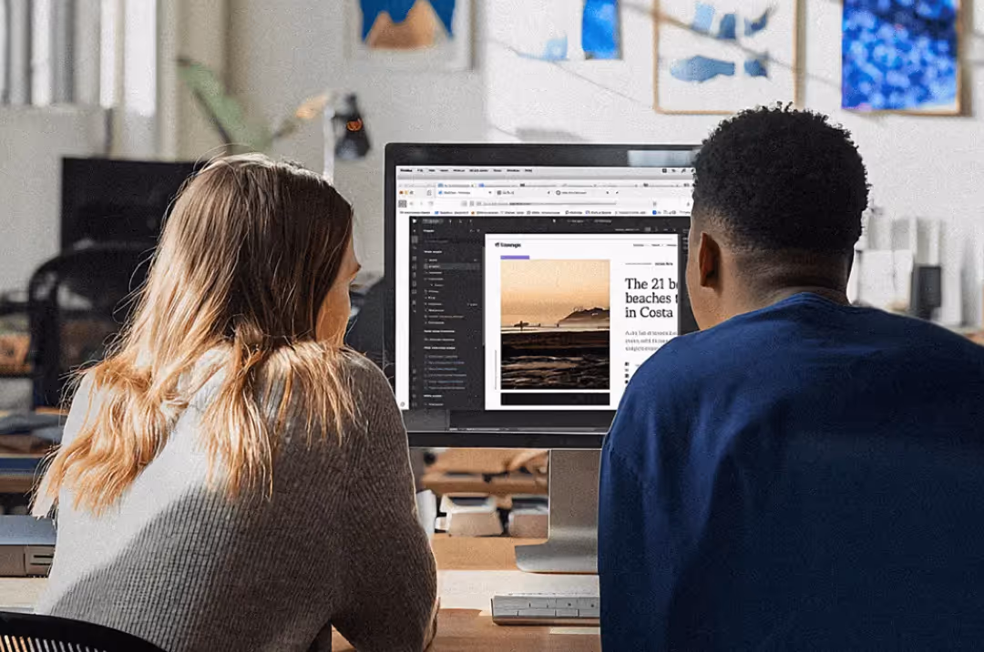 Two people sitting side by side looking at a computer screen displaying a webpage with a beach photo and article text.