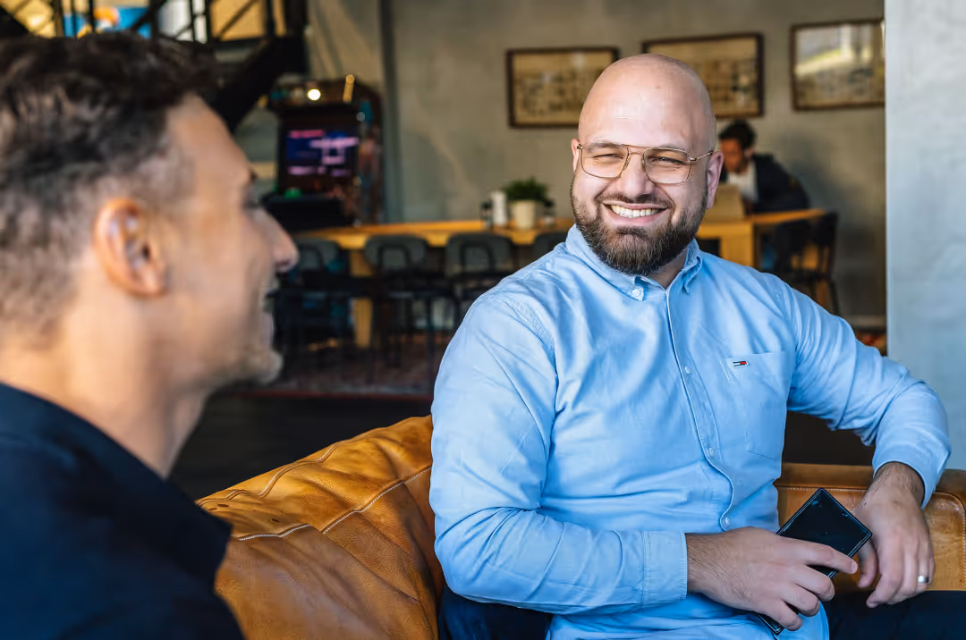 Two men sitting on a brown leather couch, one wearing glasses and a light blue shirt smiling while holding a phone.