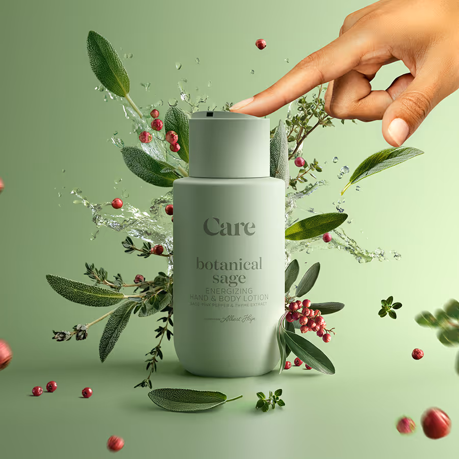 Hand pressing cap of botanical sage energizing hand and body lotion bottle surrounded by sage leaves, red peppercorns, and water splashes on light green background.