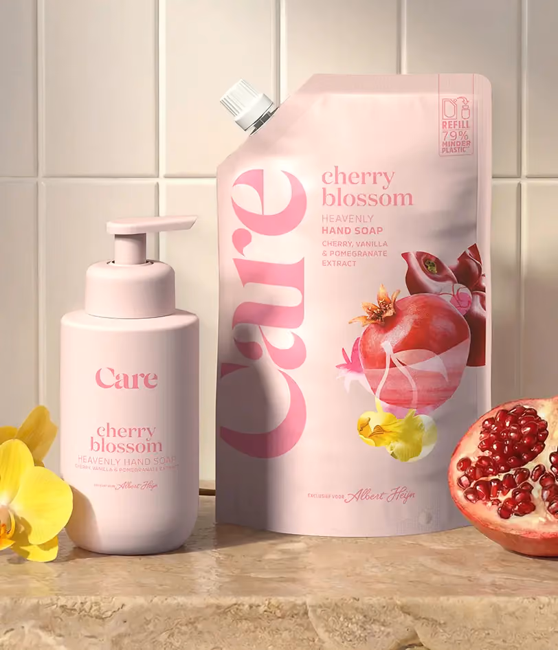Pink bottle and refill pouch of Care cherry blossom hand soap with cherry, vanilla, and pomegranate extract on tiled countertop next to yellow flowers and half a pomegranate.