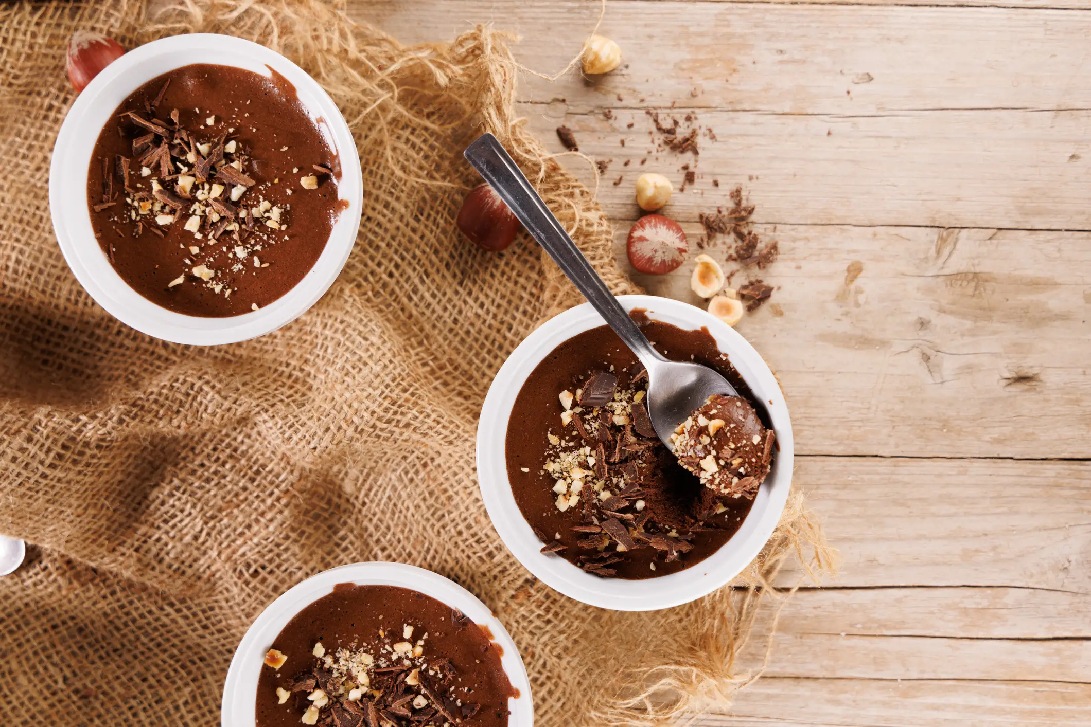 Three bowls of chocolate mousse topped with chocolate shavings and chopped nuts on a burlap cloth over a wooden surface, with a spoon in one bowl.