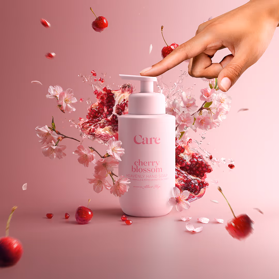 Hand pressing a pump bottle of Care cherry blossom hand soap surrounded by cherries, cherry blossoms, and pomegranate seeds splash on a pink background.