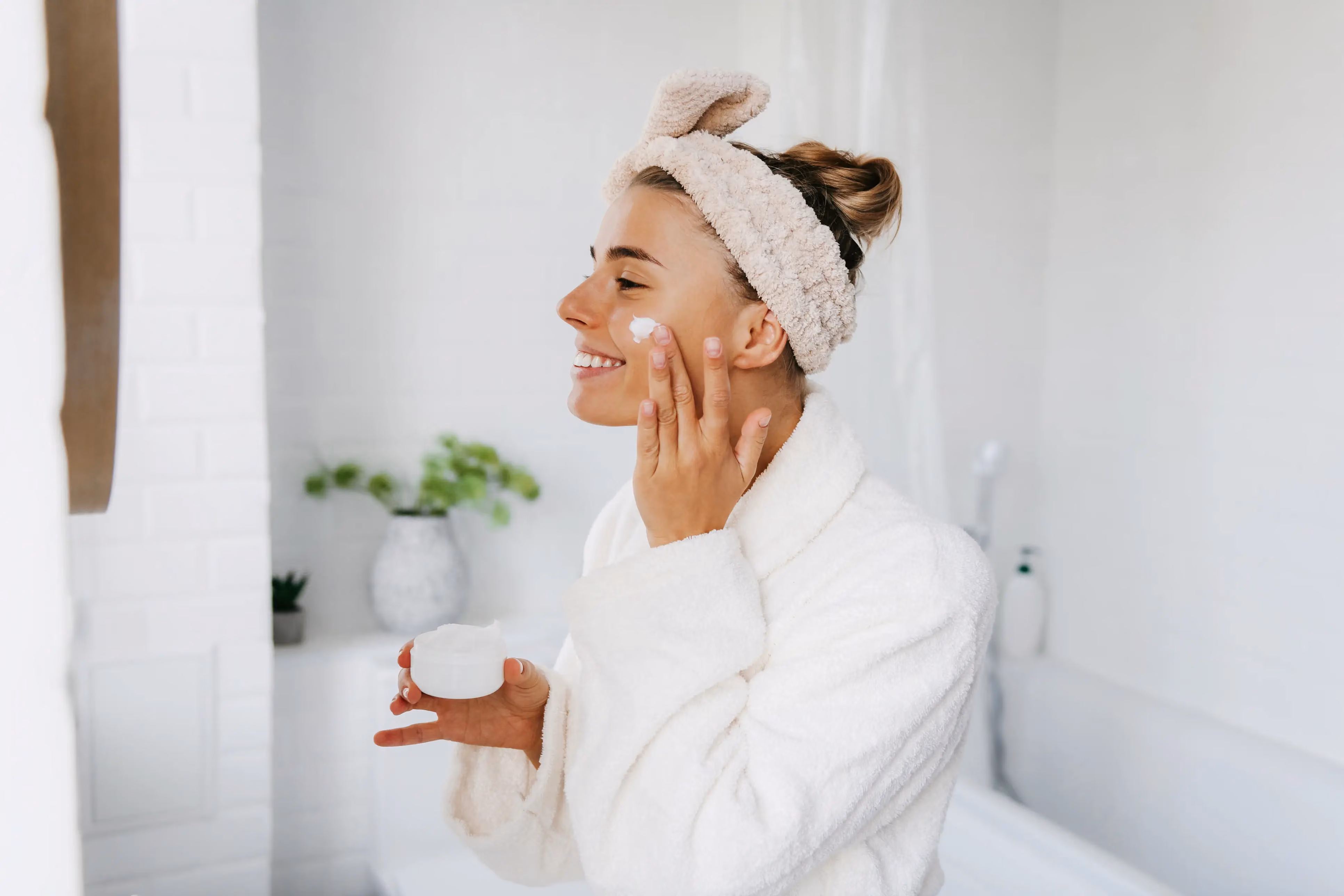 Smiling woman in a white bathrobe applying face cream in a bathroom with a headband holding her hair back.