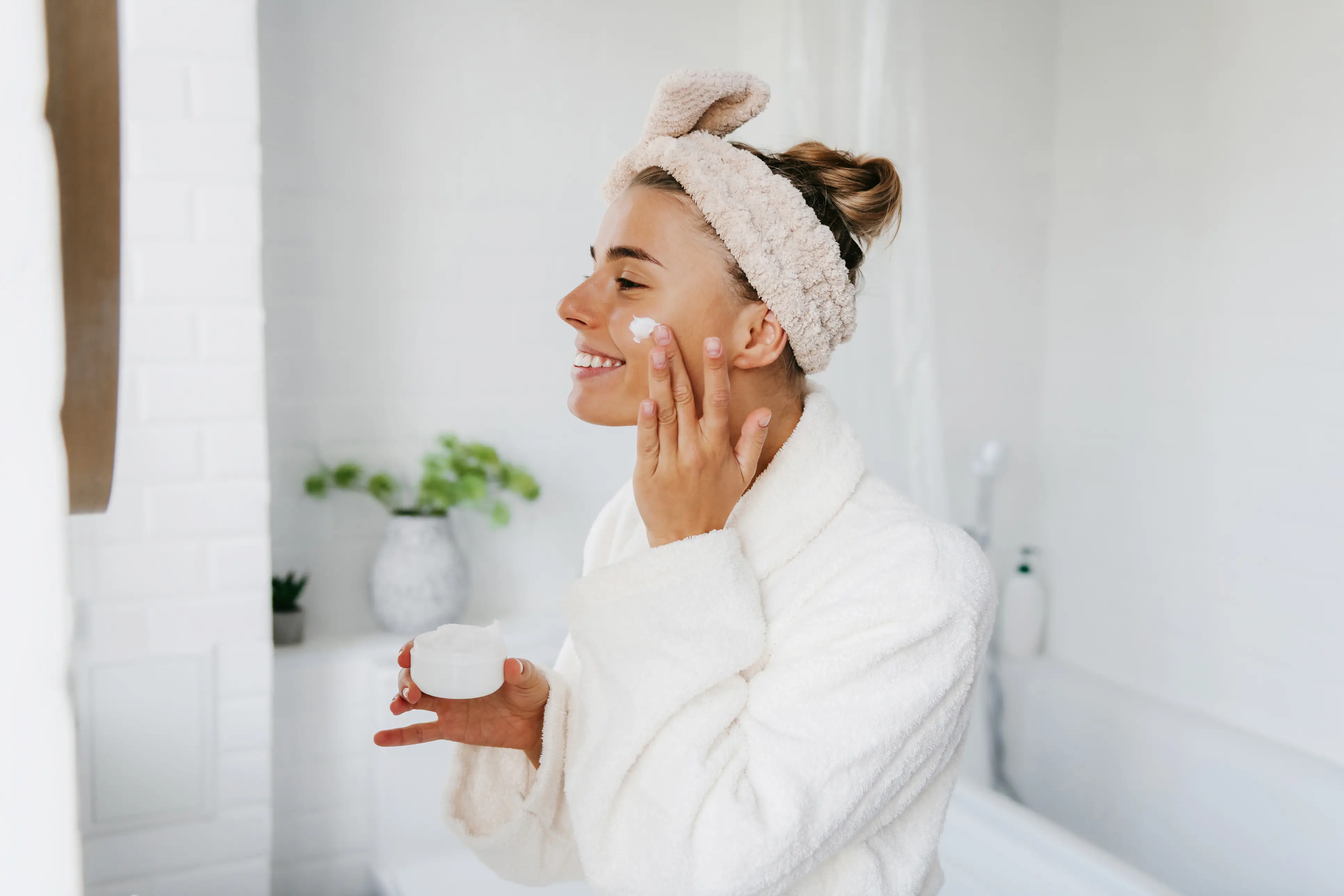 Smiling woman in a white bathrobe applying face cream in a bathroom with a headband holding her hair back.