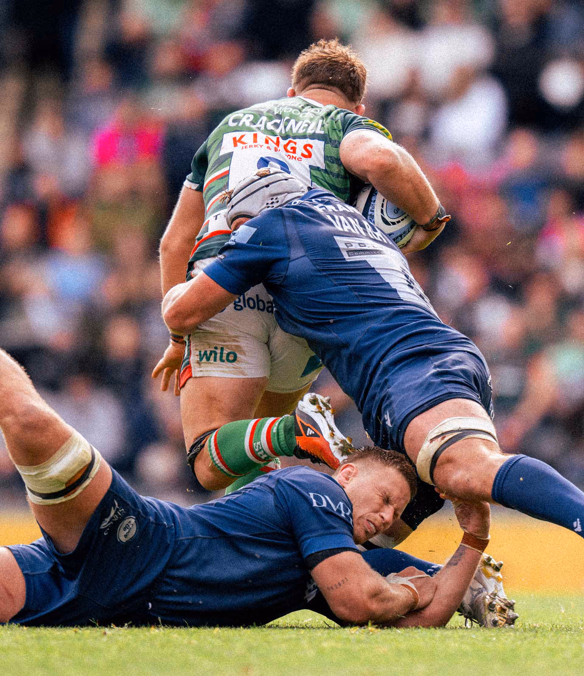 Rugby player in a green and red jersey holding the ball while tackled by two players in blue jerseys.