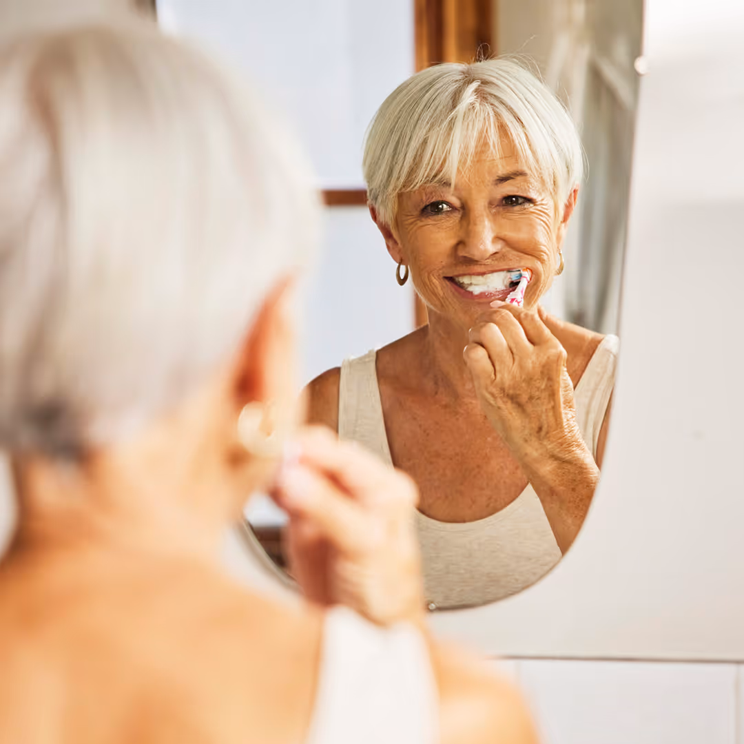 Smiling elderly woman brushing her teeth while looking in a bathroom mirror.
