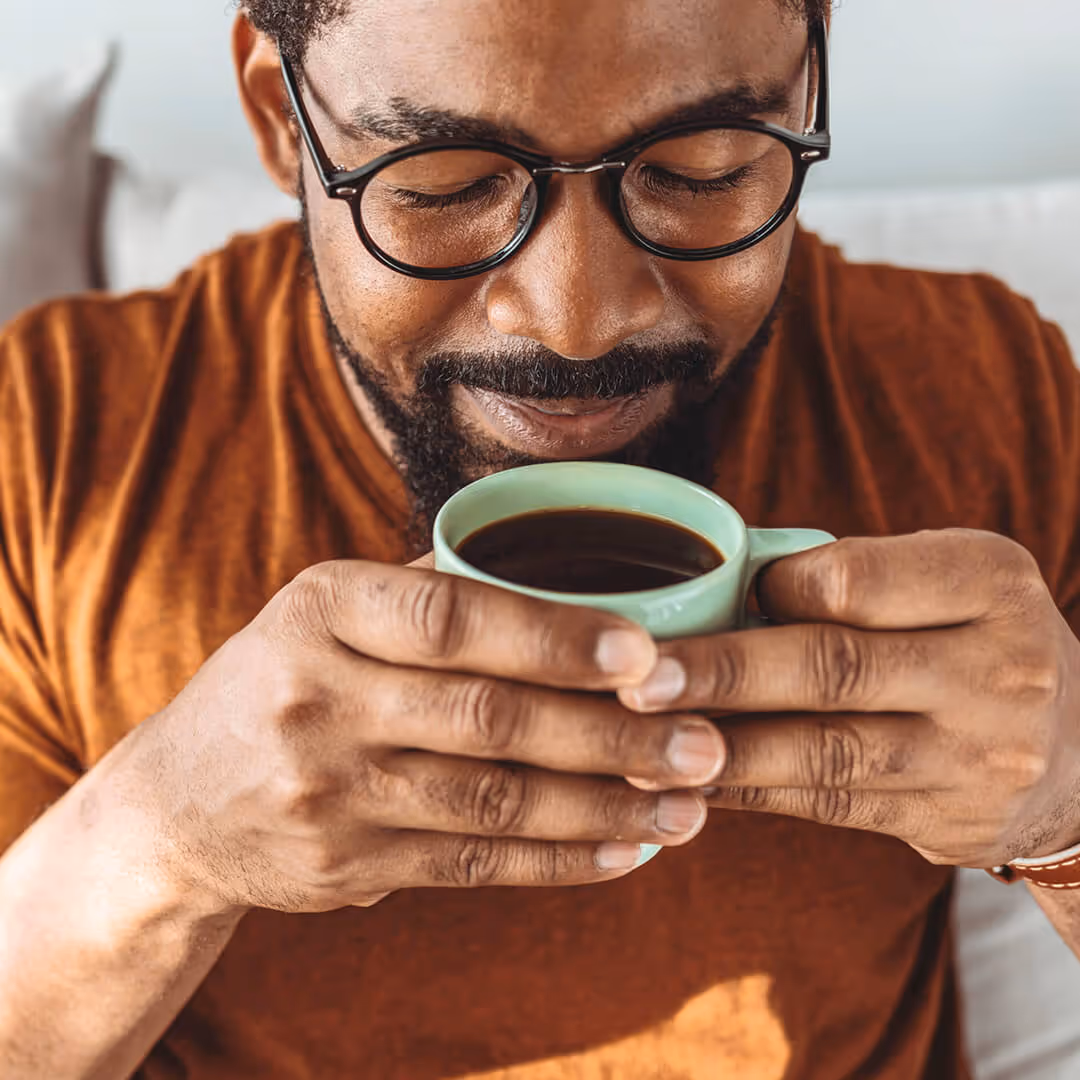 Smiling man with glasses holding and smelling a cup of coffee.