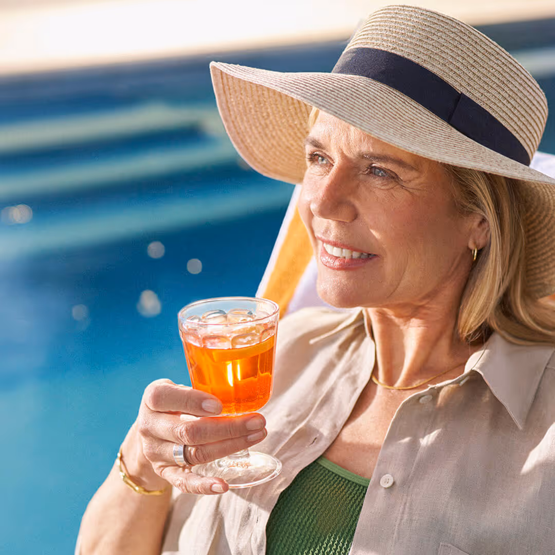 Smiling woman wearing a wide-brimmed hat holding a glass of iced orange drink by a pool.