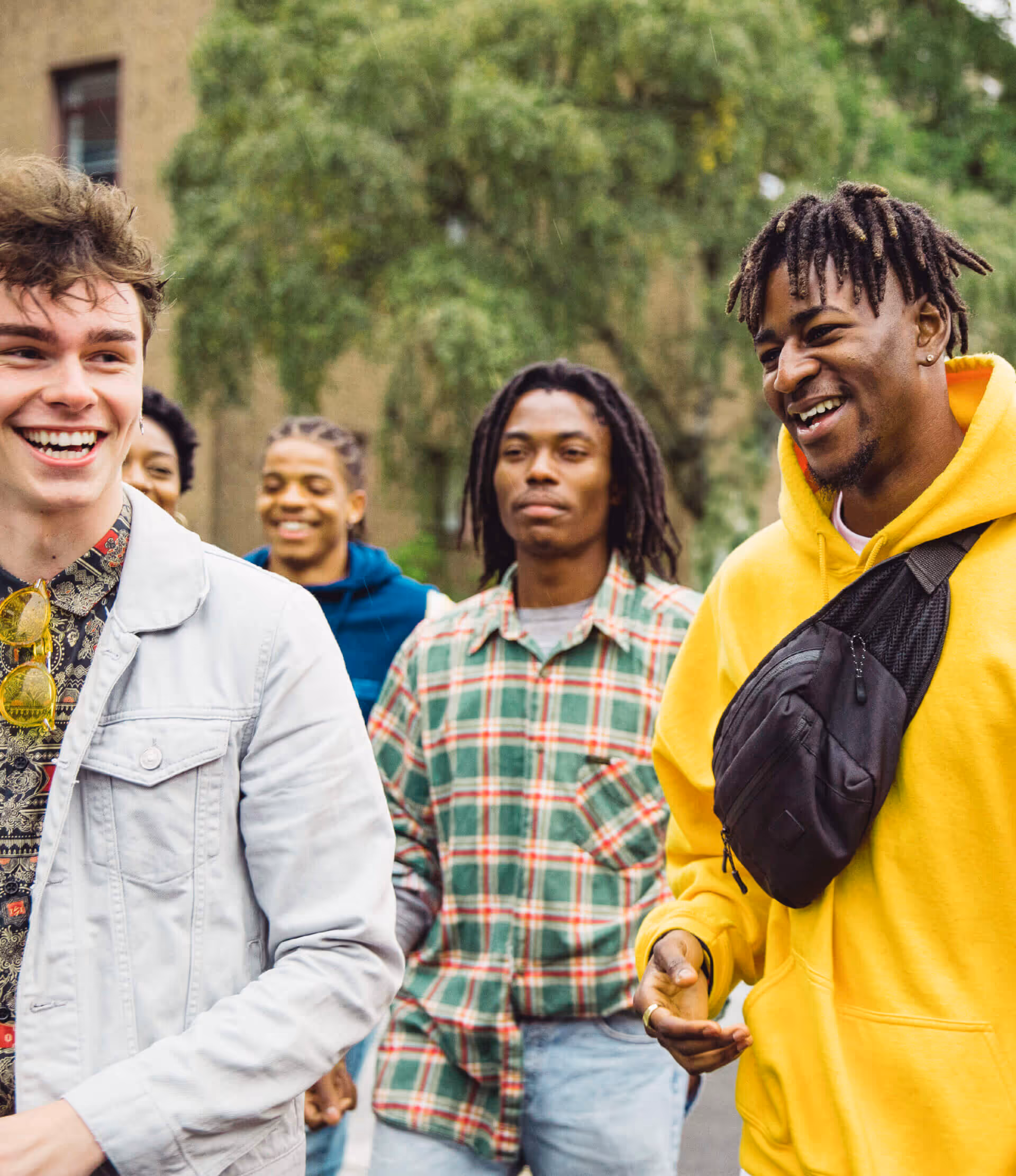 Group of five young men outdoors smiling and walking together, with trees and a building in the background.