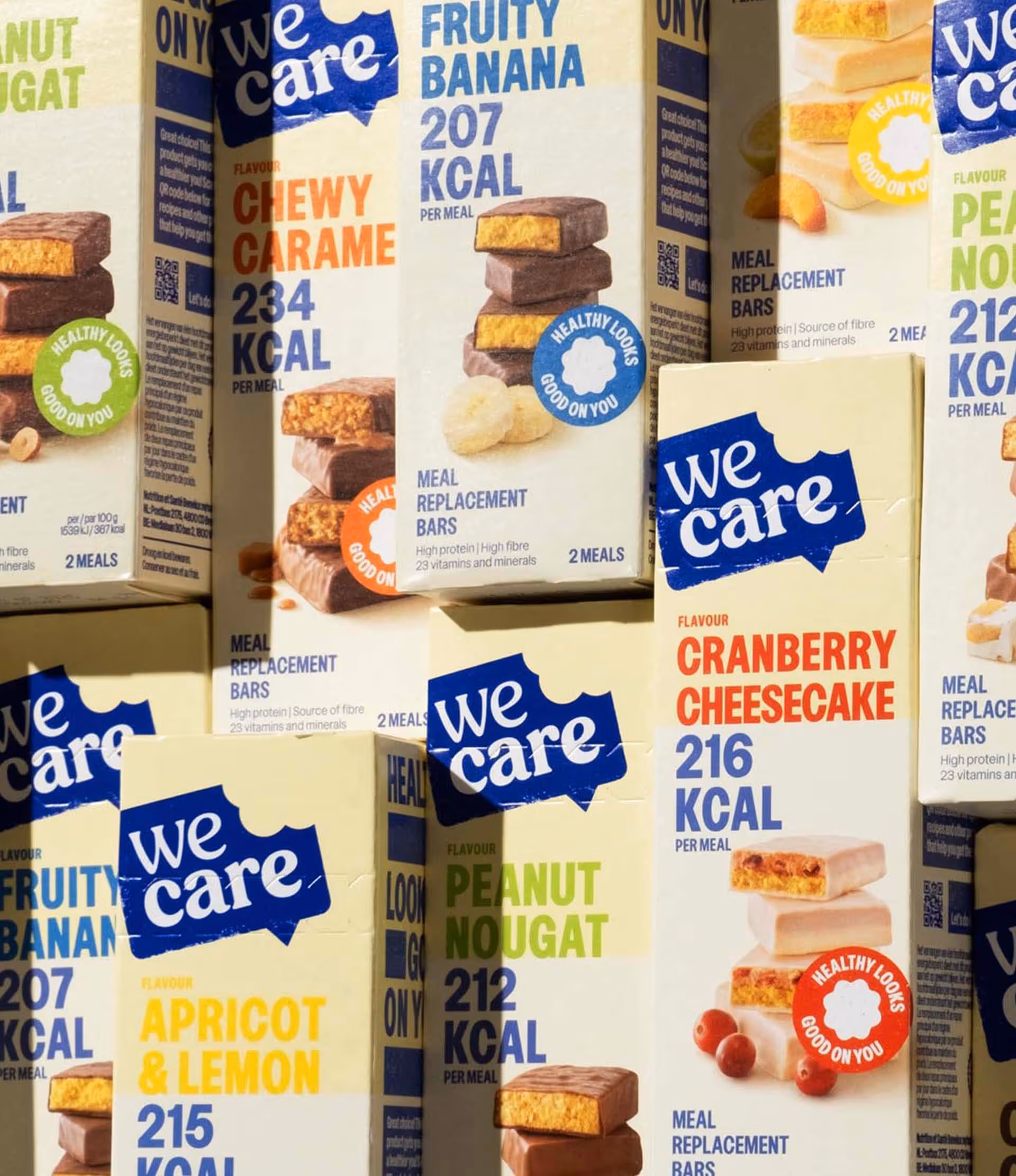 Boxes of We Care meal replacement bars in flavors like chewy caramel, fruity banana, cranberry cheesecake, peanut nougat, and apricot & lemon with calorie counts per meal.