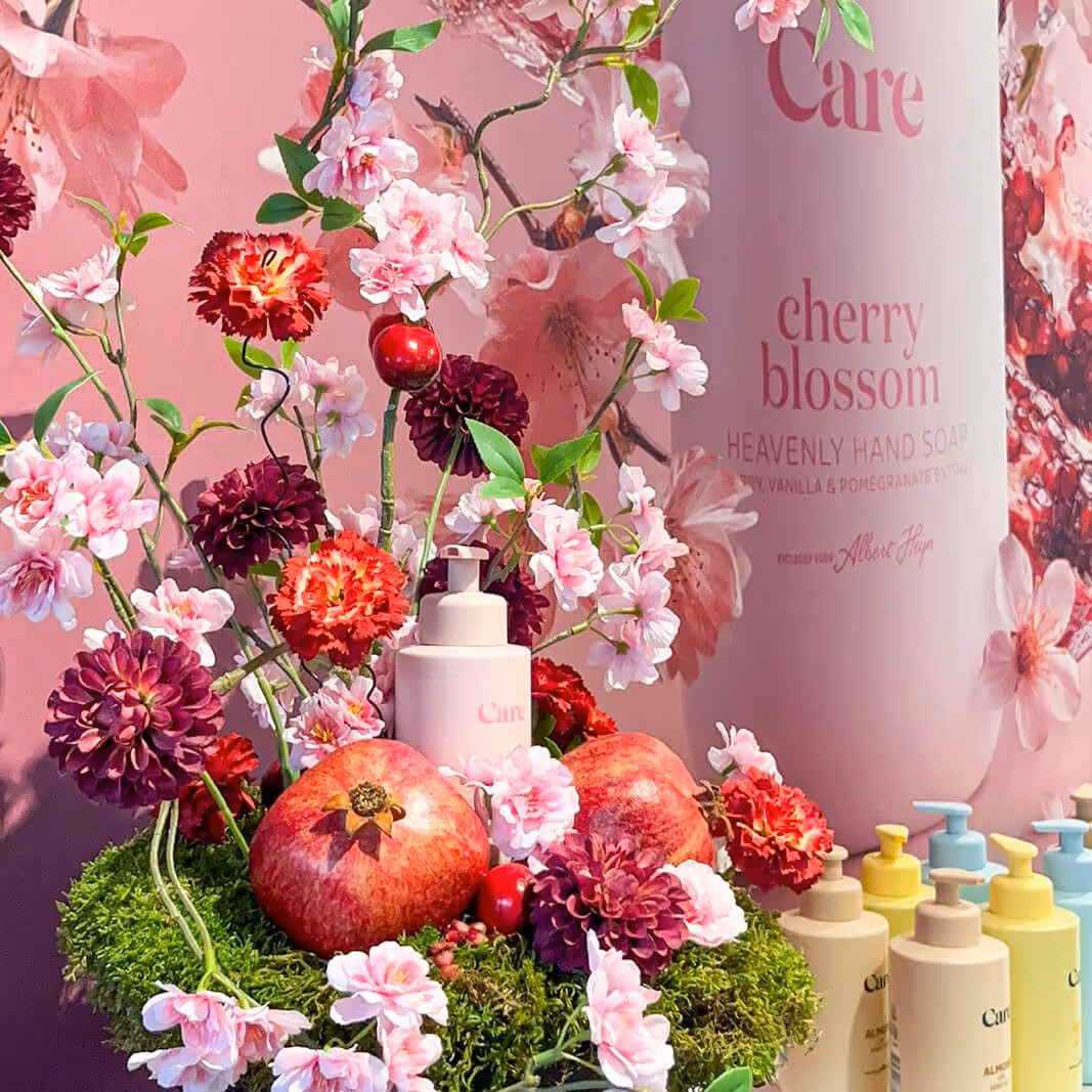 Cherry blossom hand soap bottles surrounded by pomegranates, various flowers, and green moss against a pink floral backdrop.