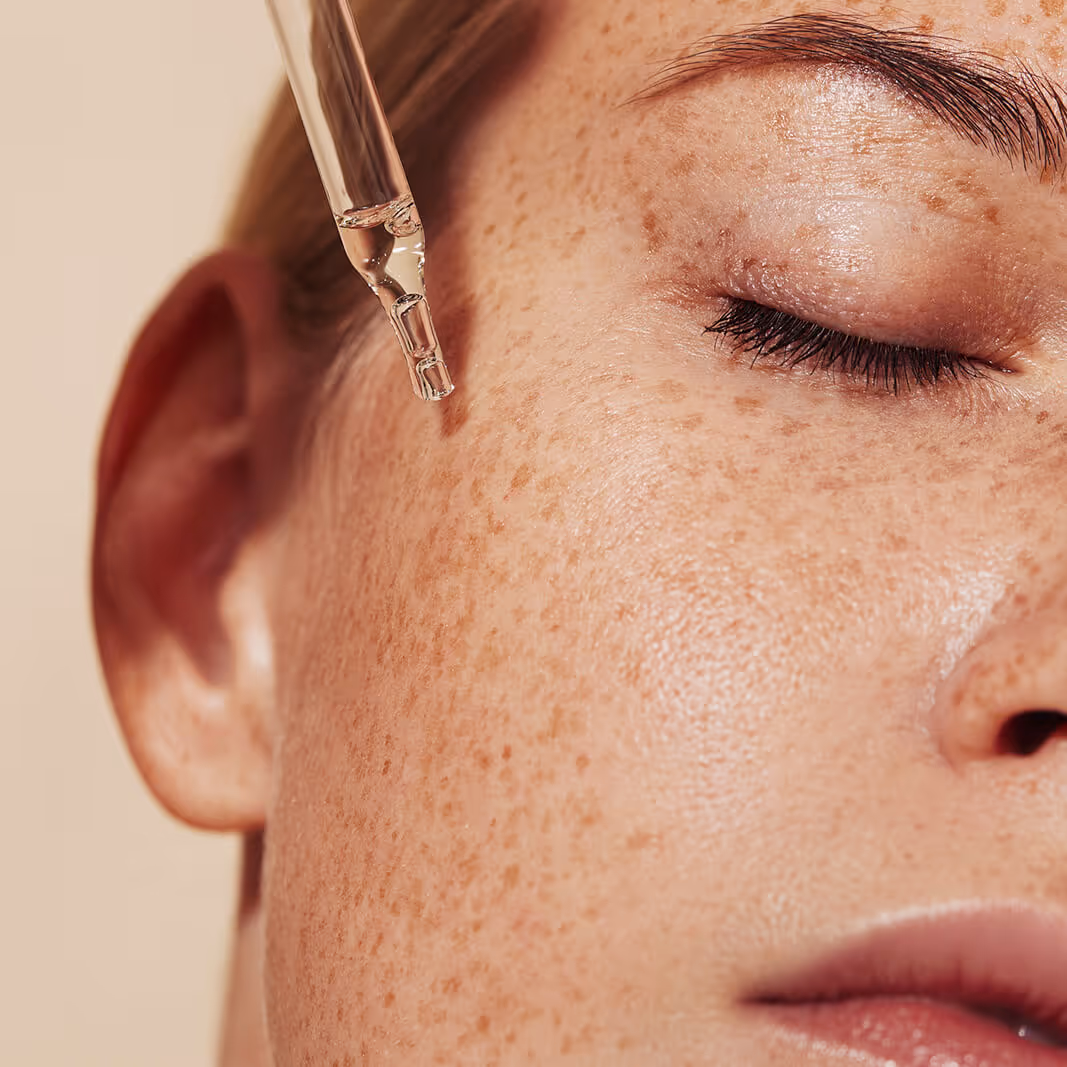 Close-up of a freckled woman's face with closed eyes as a dropper applies clear serum near her cheek.