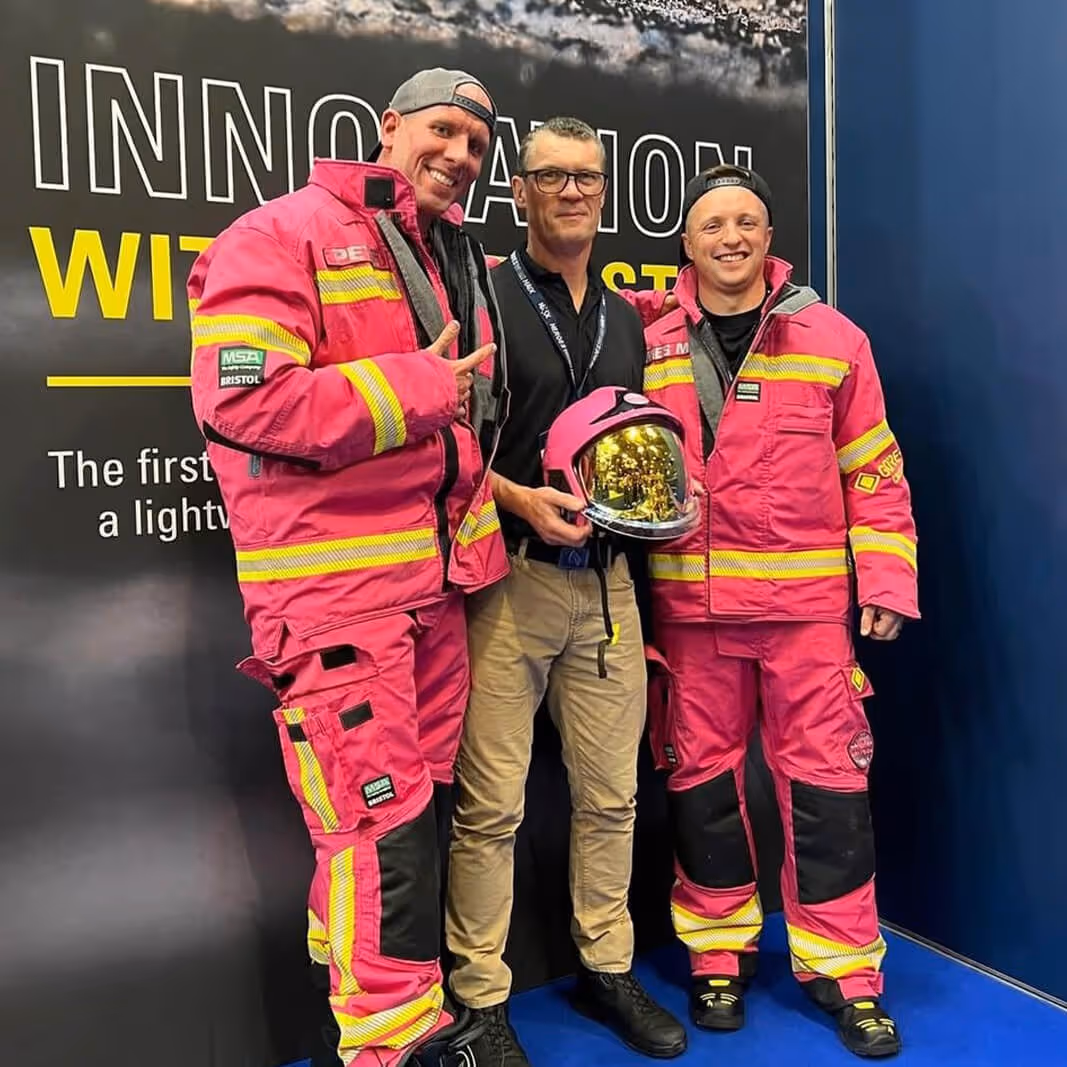 Two men in bright pink firefighting suits standing beside a man in casual clothes holding a reflective helmet, all smiling.