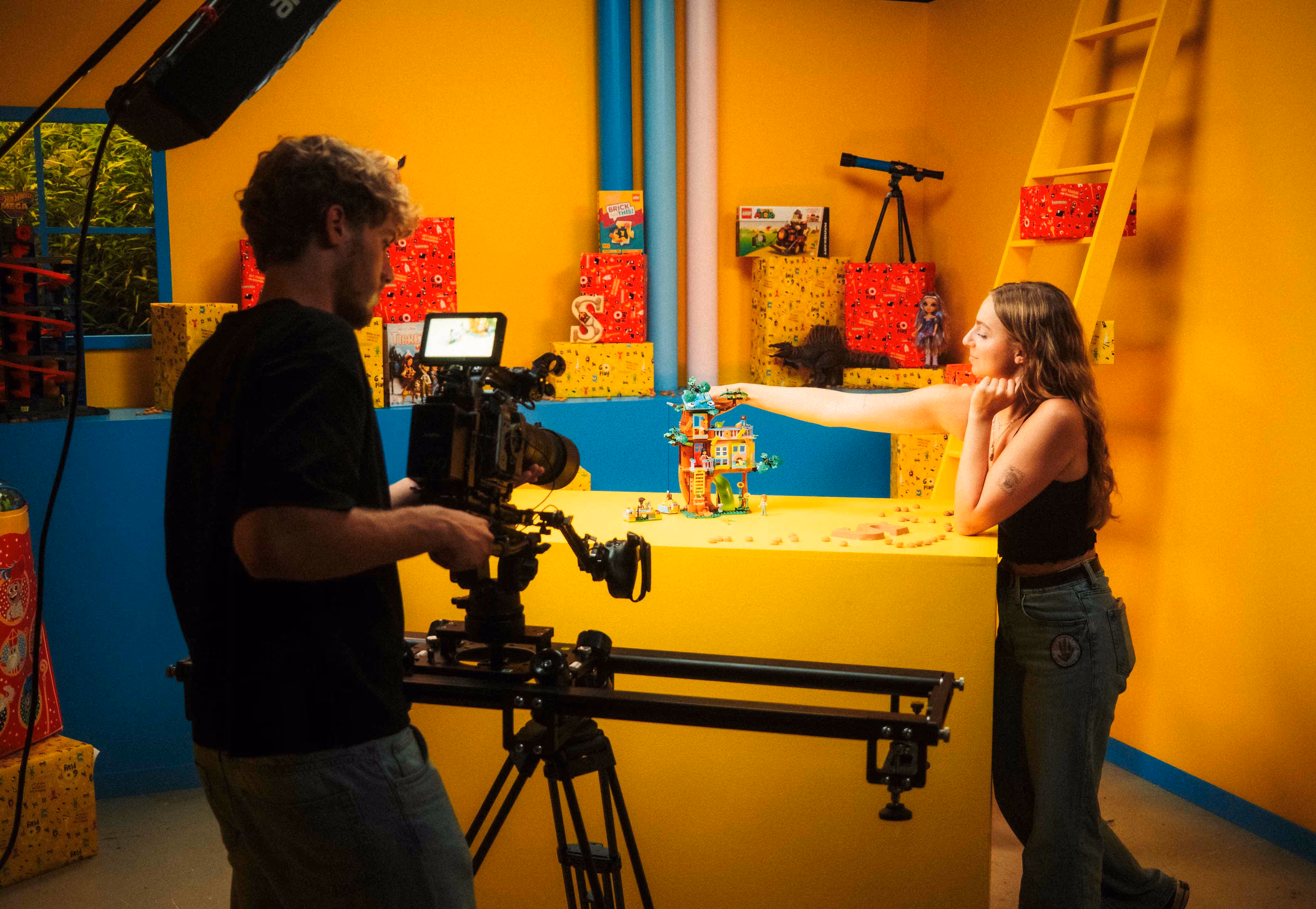 Cameraman filming a woman who is arranging a toy house on a yellow table in a brightly colored studio.