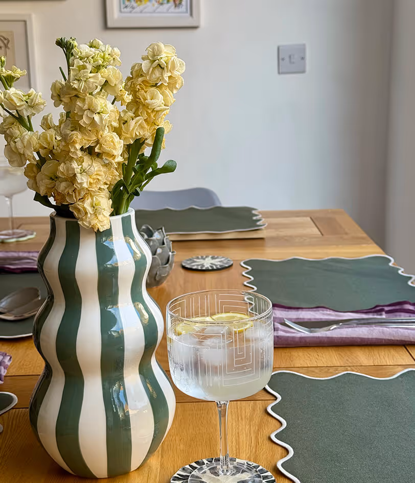 Green and white striped vase with yellow flowers on a wooden table set with green placemats and a glass of lemon water with ice.