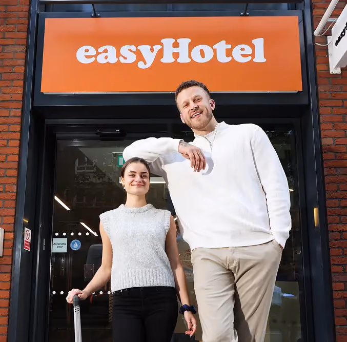 Smiling man and woman standing in front of an easyHotel entrance, the woman holding a suitcase handle.