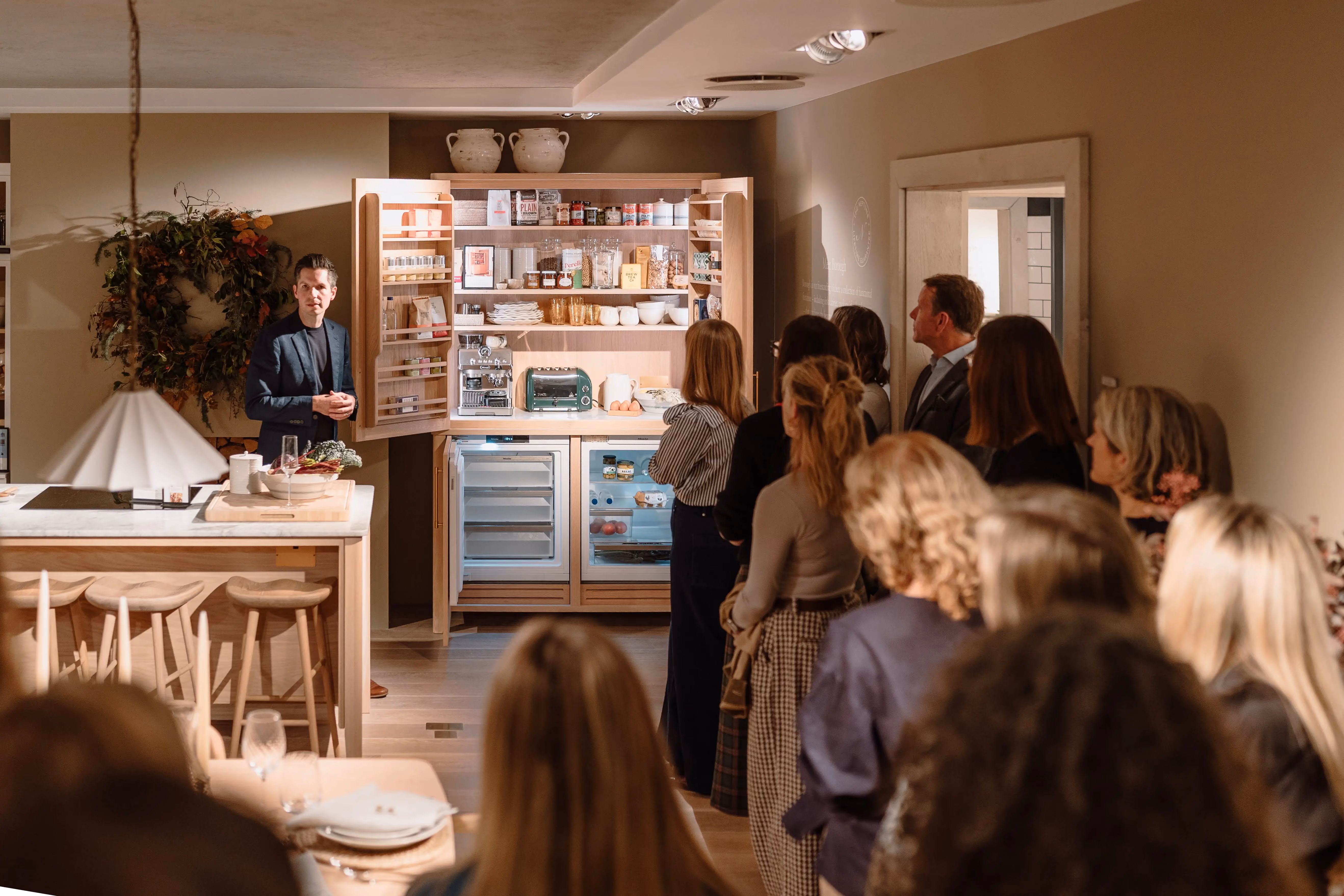 Man presenting a kitchen setup with open cupboards and appliances to an audience in a warmly lit room.