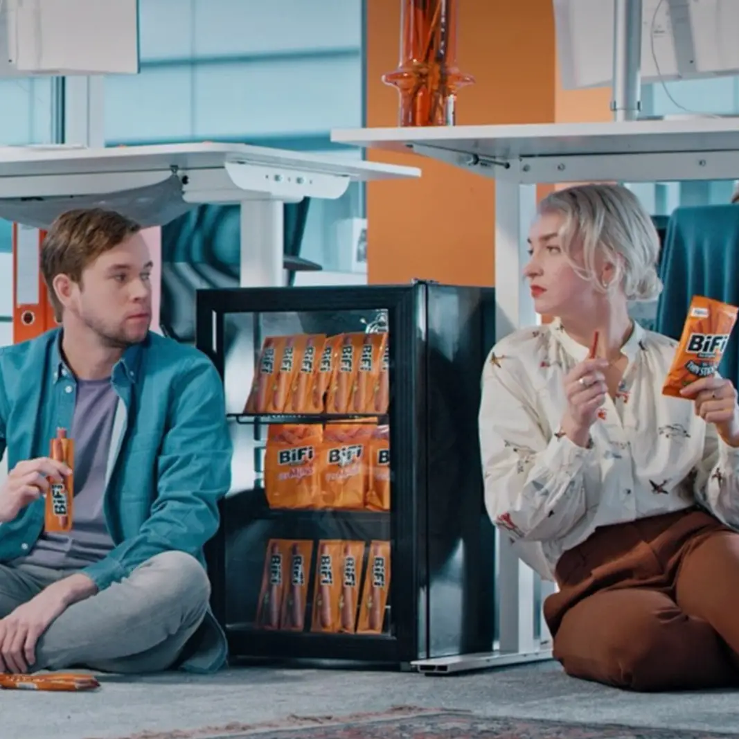 A man and a woman sitting on the floor near a small fridge filled with BiFi snack packs, each holding a BiFi snack.