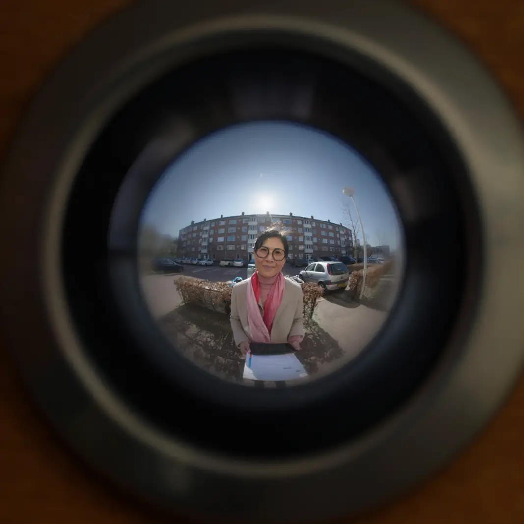 Woman with glasses and a pink scarf standing outdoors, seen through a fisheye peephole lens with a residential building and parked cars in the background.