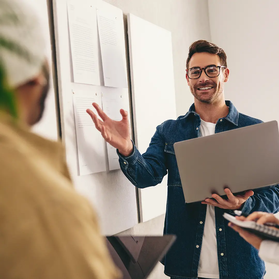 Man wearing glasses and denim jacket holding a laptop and gesturing while speaking in a meeting.