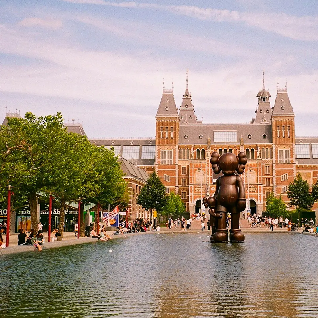 Bronze sculpture standing in a pond in front of the Rijksmuseum building with visitors sitting and walking around on a sunny day.