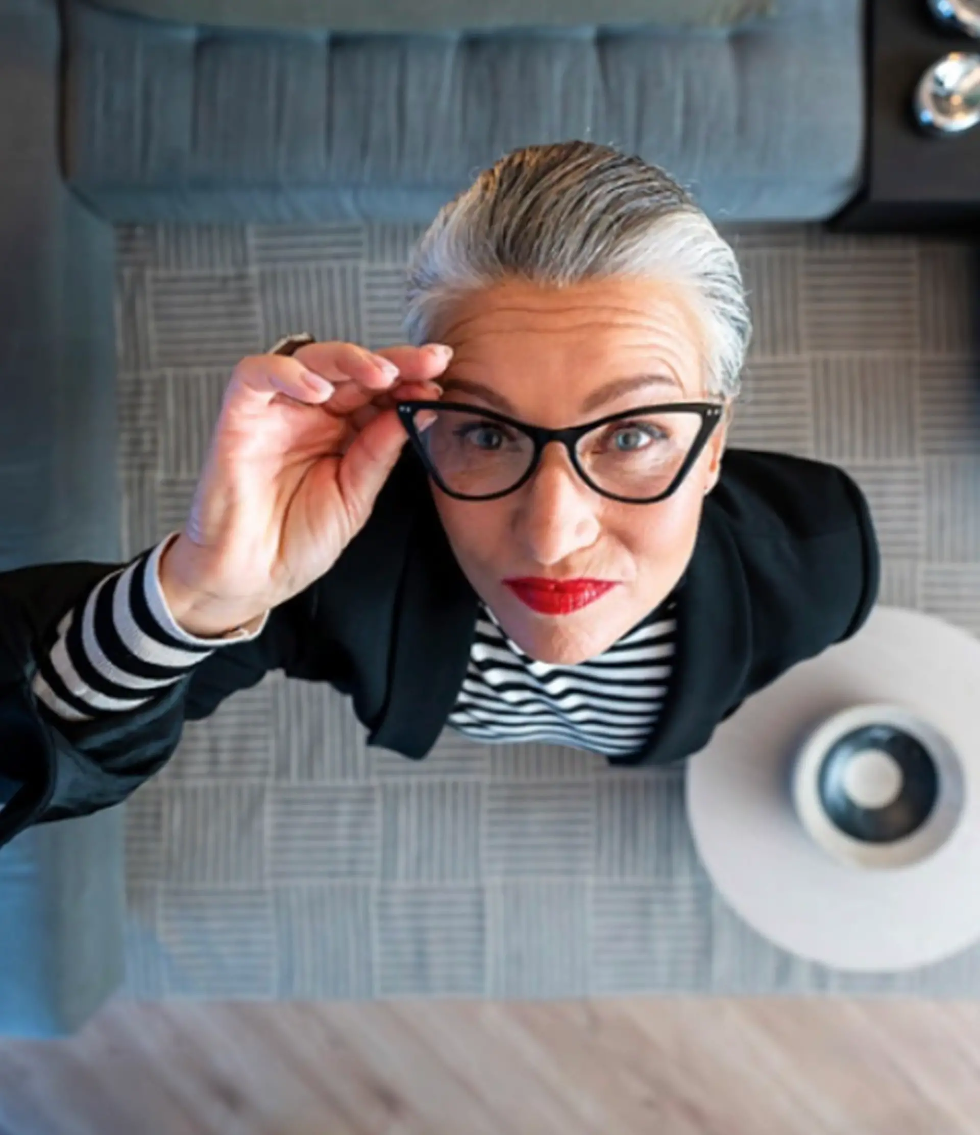 Top view of a confident older woman with gray hair, red lipstick, black cat-eye glasses, and a striped shirt under a black blazer looking upward.