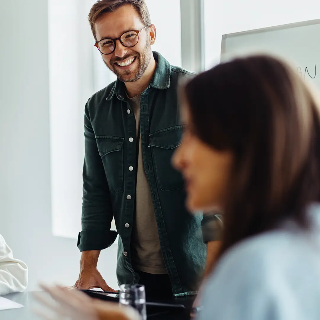 Smiling man wearing glasses and a green jacket standing in a bright room with a blurred person in the foreground.