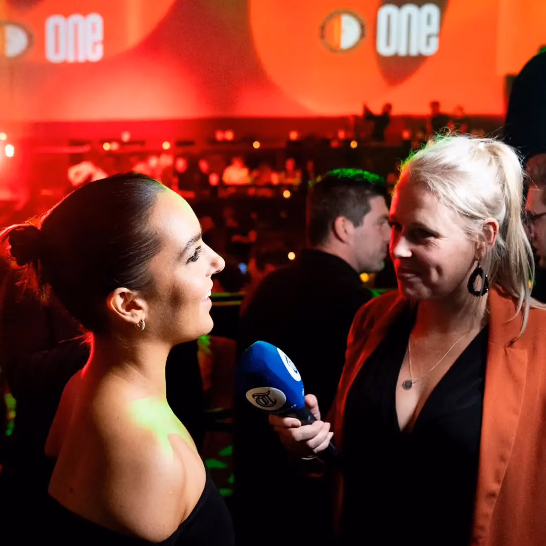 A female reporter holding a microphone interviewing a smiling woman with a bun in a dimly lit event space.