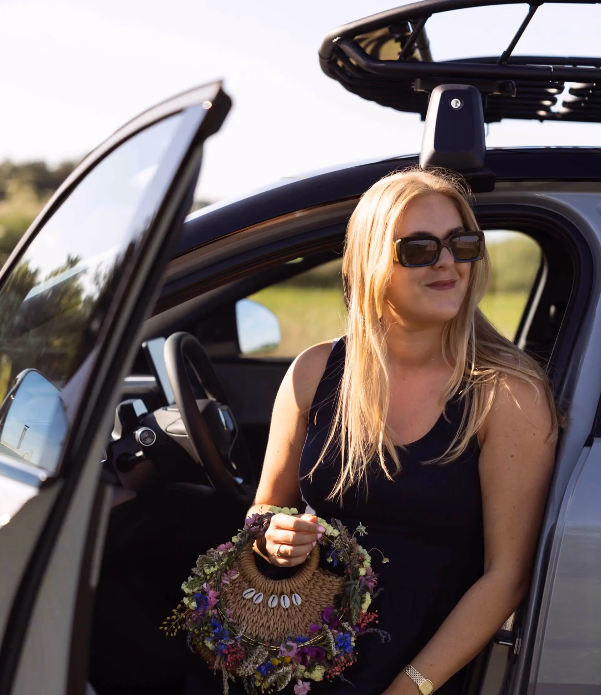 Woman with long blonde hair and sunglasses sitting in an open car door holding a decorative floral wreath bag.
