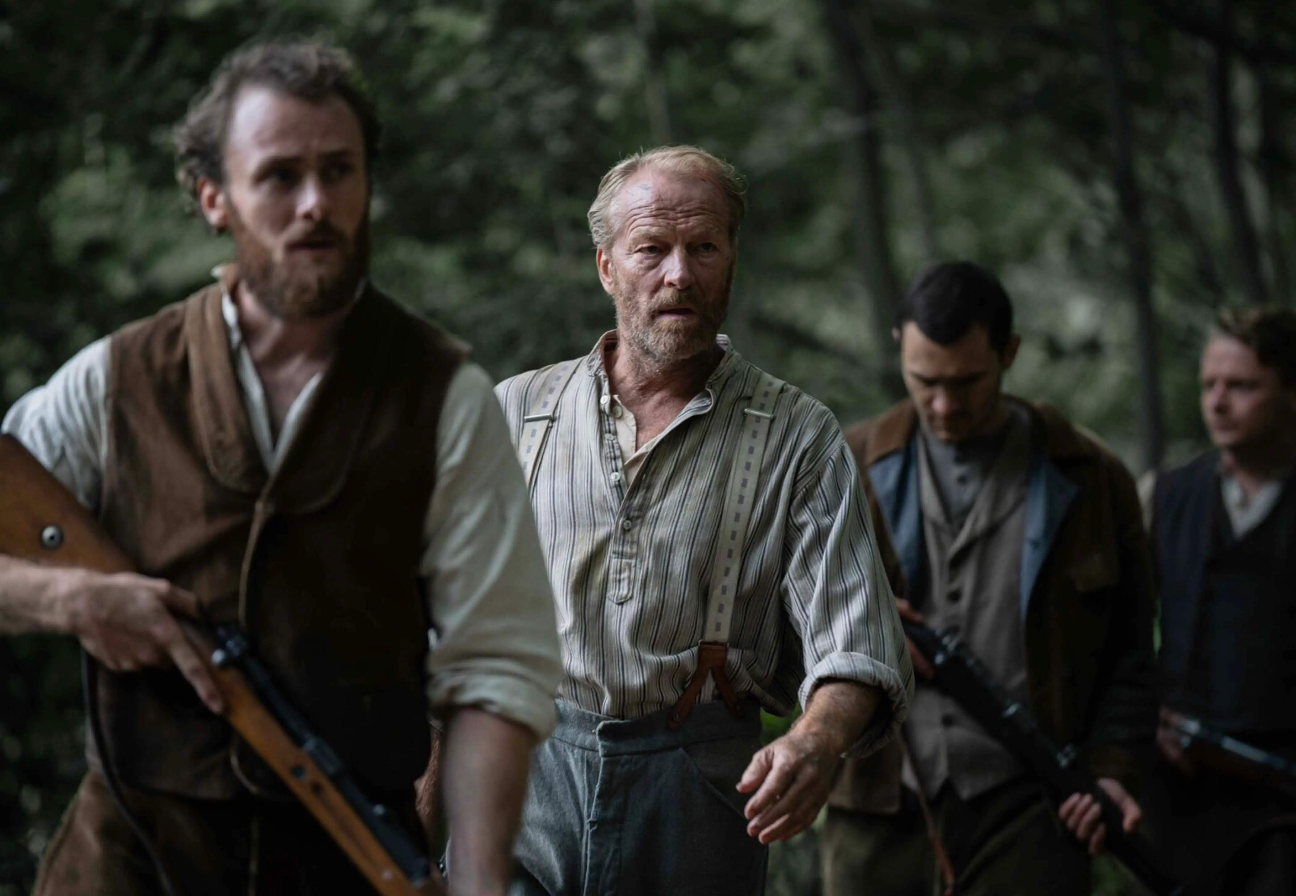 Four men with serious expressions walking through a forest, two holding rifles.