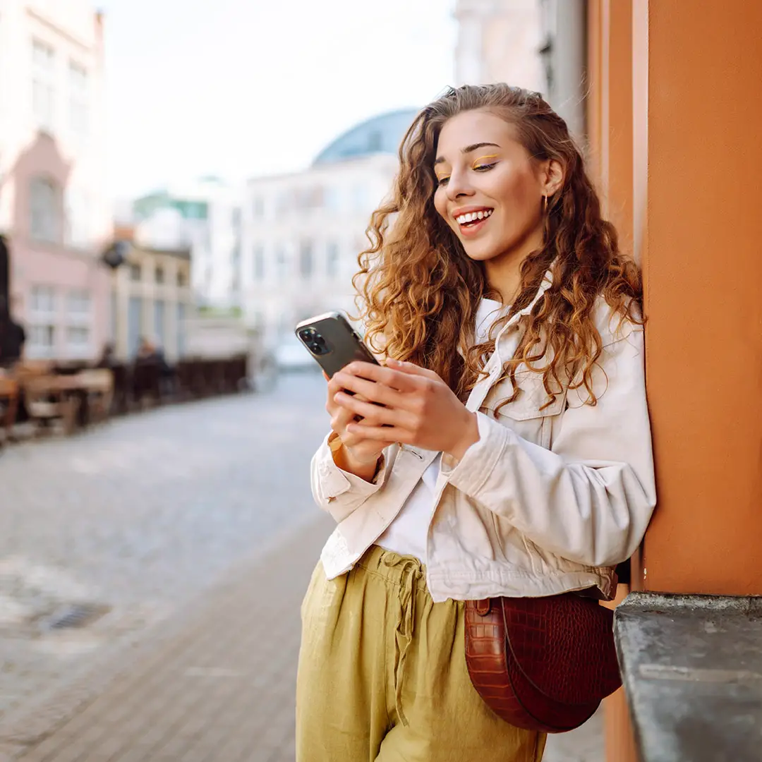 Smiling woman with curly hair leaning against a building while looking at her smartphone on a city street.