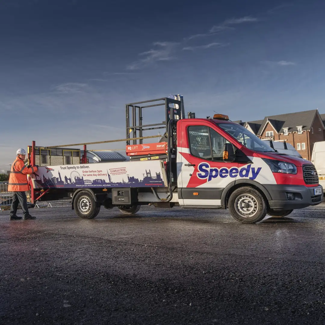 Red and white Speedy delivery truck with a worker in an orange safety jacket and white helmet standing beside it on a paved area under a blue sky.