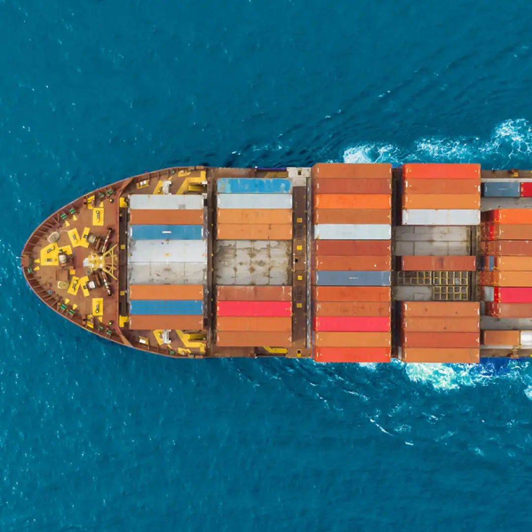 Aerial view of the bow of a cargo ship loaded with multicolored shipping containers sailing in blue ocean water.