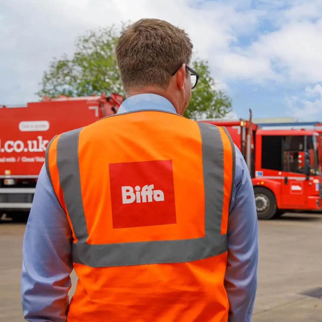 Man wearing an orange high-visibility vest with 'Biffa' logo stands in front of red Biffa trucks outdoors.