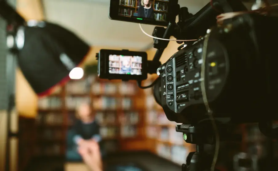 Video camera focused on recording a person sitting in a library setting with bookshelves in the background.
