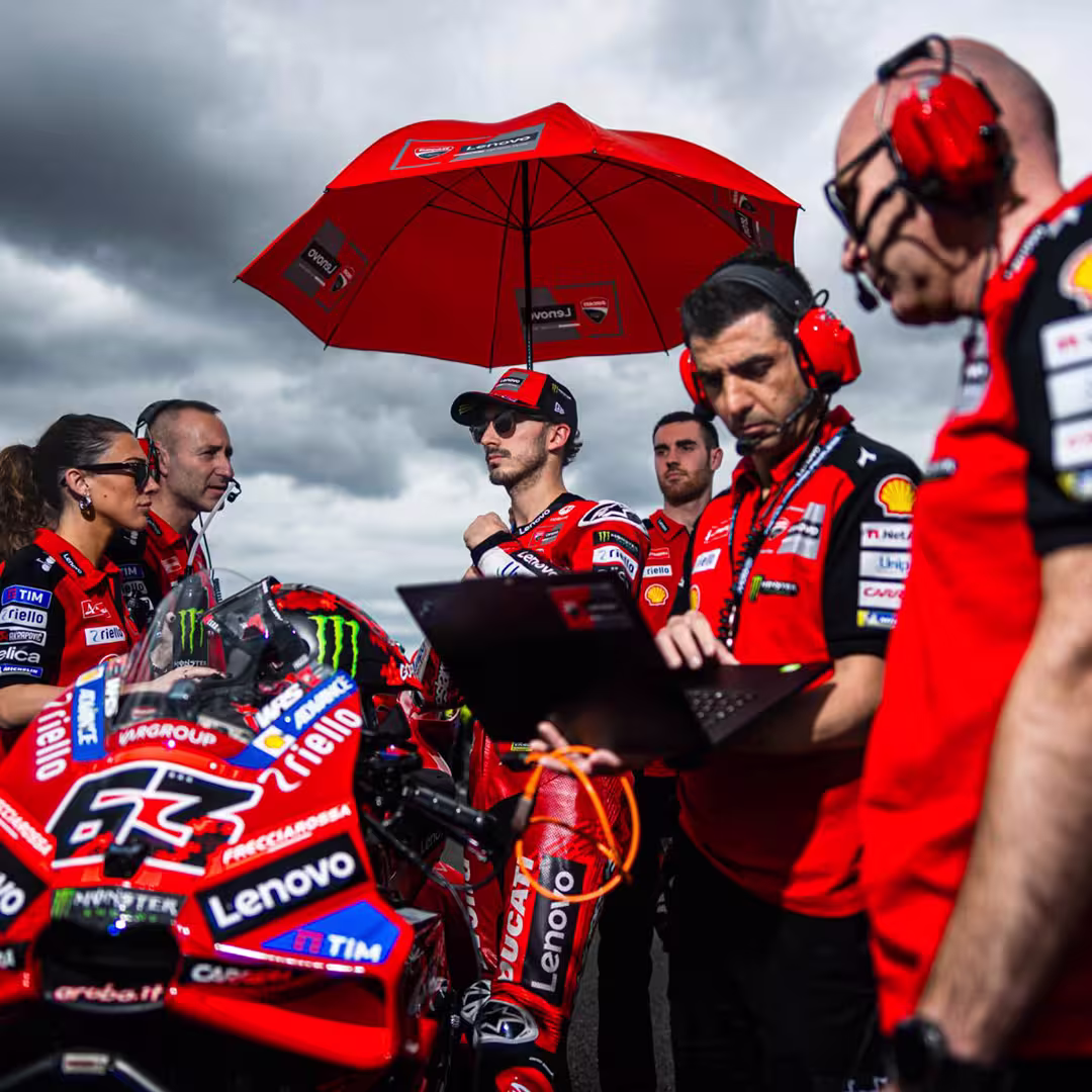 Motorcycle racer in red gear standing next to a red Ducati racing bike under a branded red umbrella with team members around.
