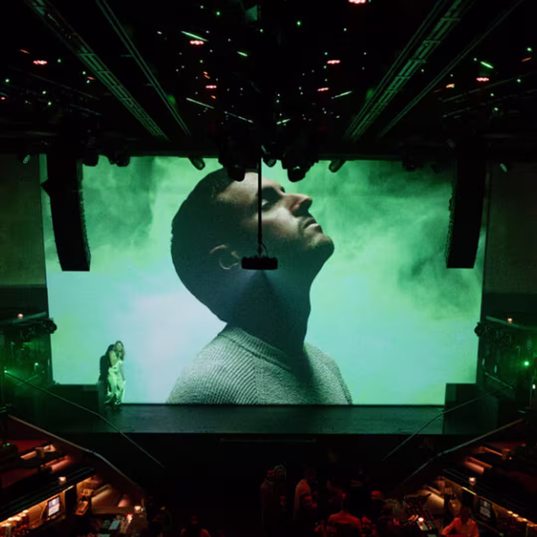 Large stage screen displaying a close-up profile of a man looking upwards with a green smoky background, in a dimly lit theater with people and seating below.