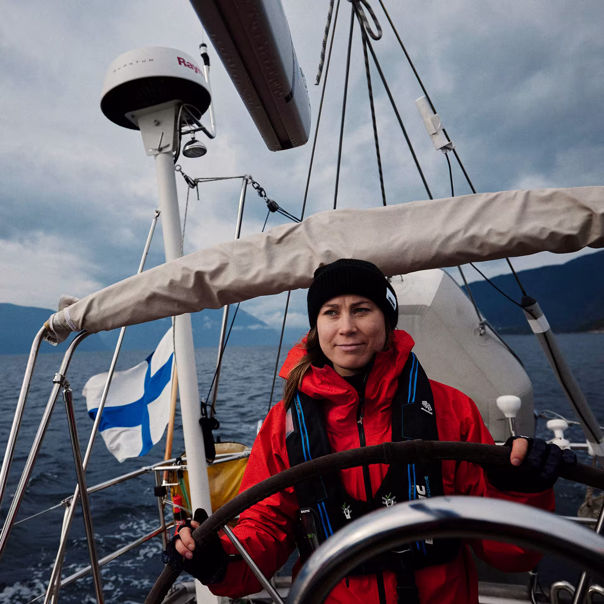 A woman in a red sailing jacket and black beanie at the helm of a sailboat, with a Finnish flag flying and a Raymarine radar dome mounted on the mast, against a backdrop of dark mountains and overcast skies.