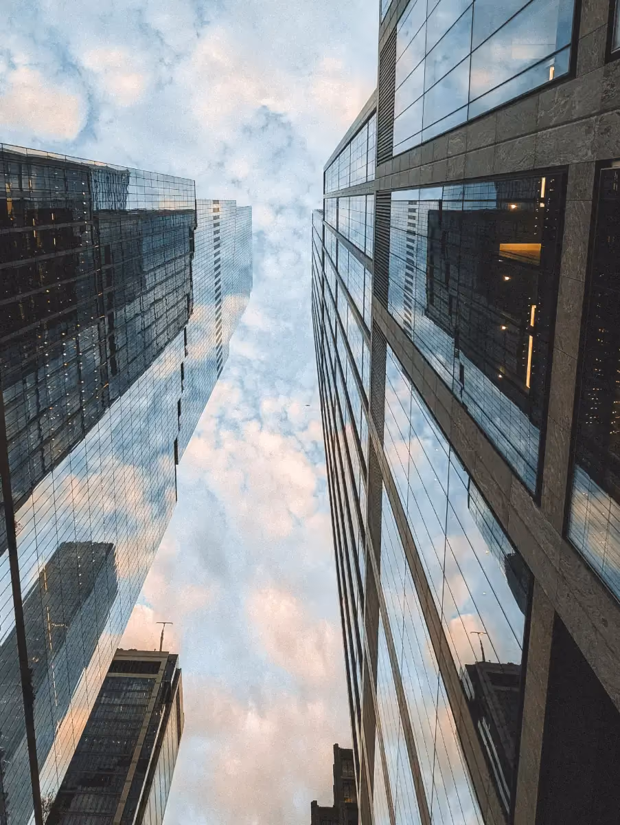 Looking up between tall glass skyscrapers reflecting a cloudy sky in an urban setting.