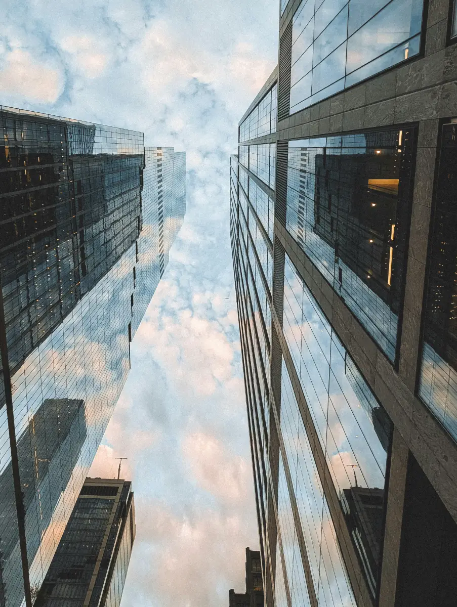 Looking up between tall glass skyscrapers reflecting a cloudy sky in an urban setting.