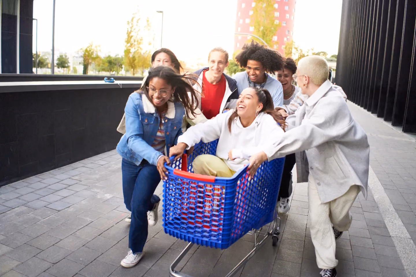 A group of friends smiling and pushing a blue shopping cart with one friend sitting inside outdoors.