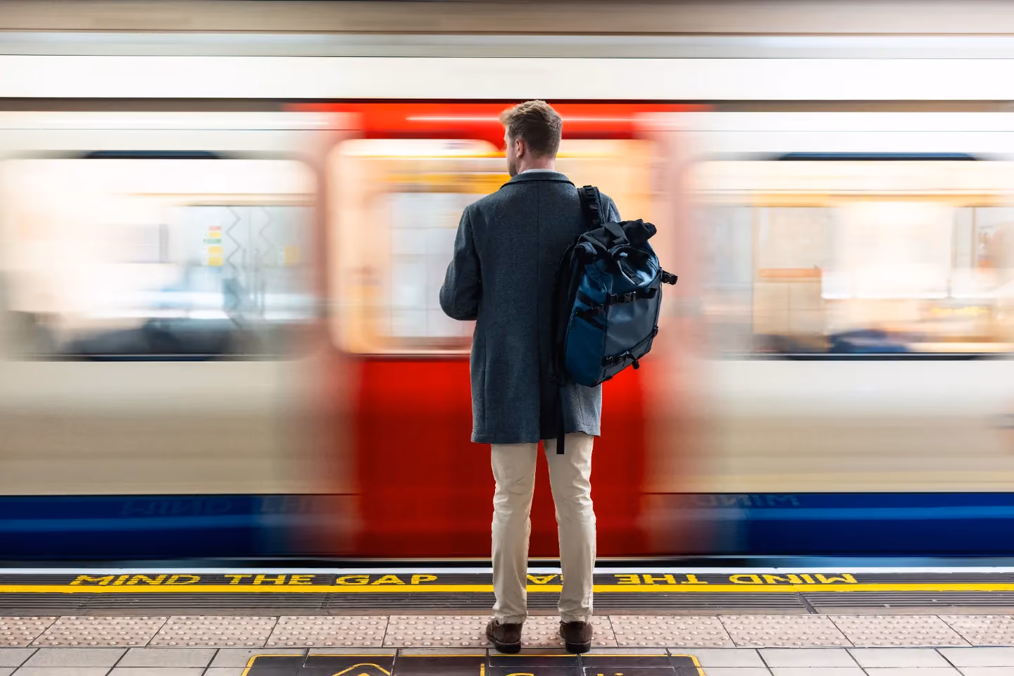 Man with backpack waiting on subway platform as train passes quickly behind him.