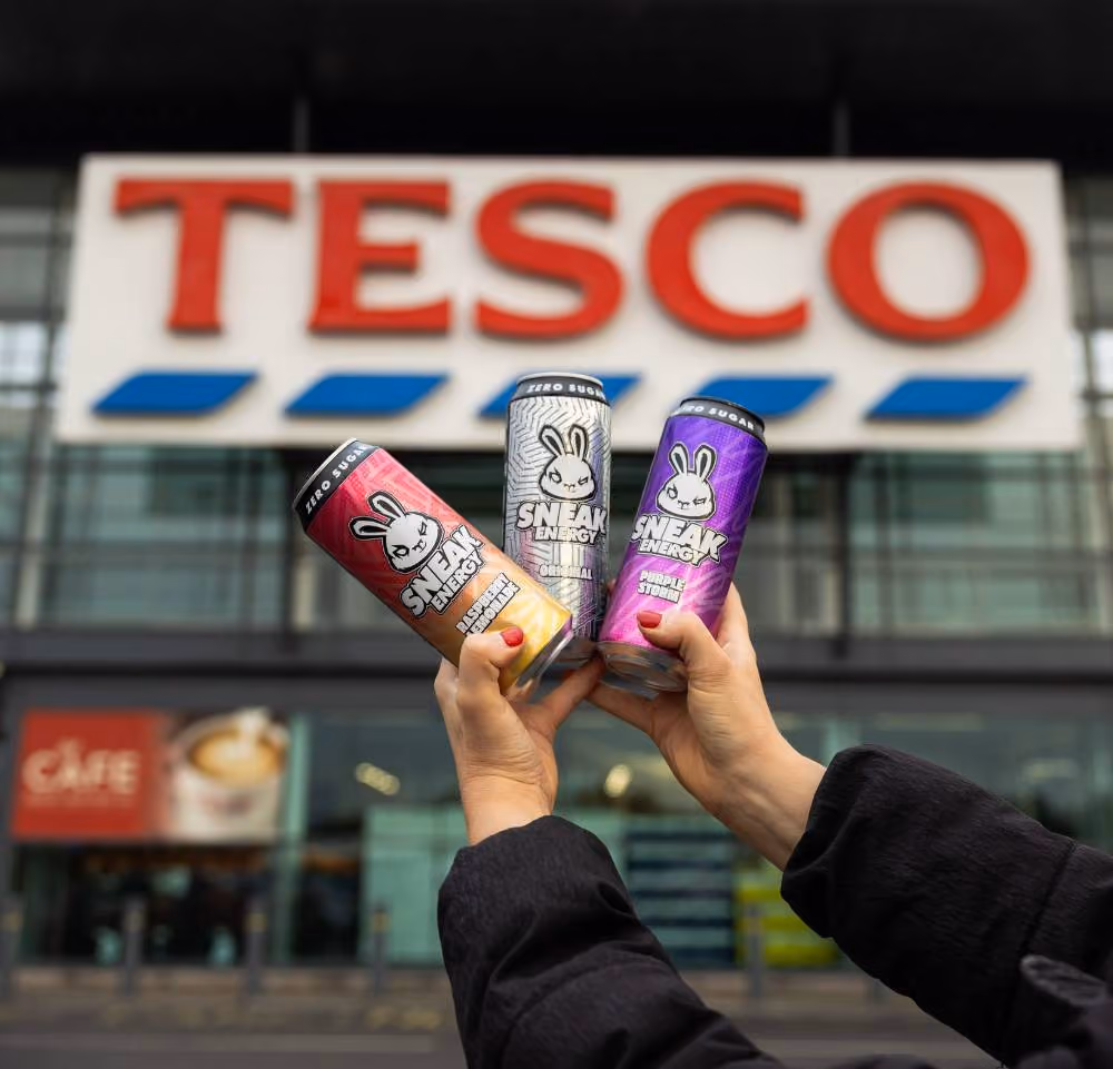 Three cans of Sneak Energy drink held in hand outside a Tesco store with the Tesco sign visible.