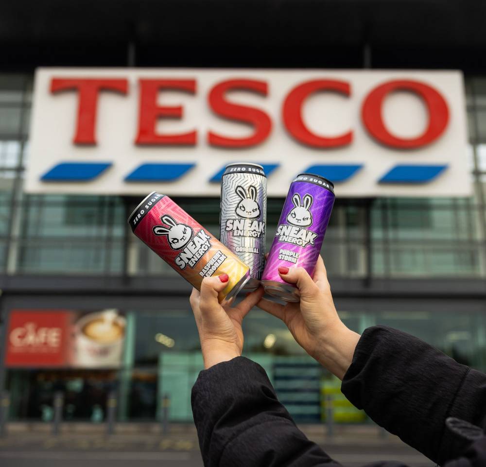Three cans of Sneak Energy drink held in hand outside a Tesco store with the Tesco sign visible.