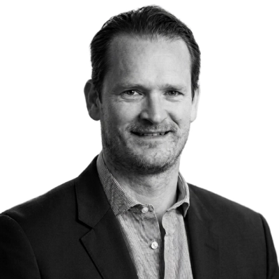 Black and white headshot of a middle-aged man with short dark hair, wearing a dark blazer over a patterned button-up shirt, smiling slightly against a white background.